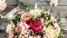 Un bouquet di fiori freschi con rose rosa, garofani bianchi e una peonia rossa, arricchito da foglie verdi, su uno sfondo di legno rustico.
