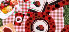Set di articoli da festa a tema coccinella, con piatti, bicchieri e tovaglioli decorati con motivi di coccinelle rosse e nere, disposti su una tovaglia a quadri rossi e bianchi, accompagnati da frutta fresca.
