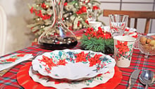 Due piatti bianchi con decorazioni floreali uno sopra l'altro sopra una tavola apparecchiata per Natale, recante una tovaglia rossa, un decanter con vino rosso e una decorazione di agrifoglio.