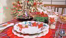 Due piatti bianchi con decorazioni floreali uno sopra l'altro sopra una tavola apparecchiata per Natale, recante una tovaglia rossa, un decanter con vino rosso e una decorazione di agrifoglio.