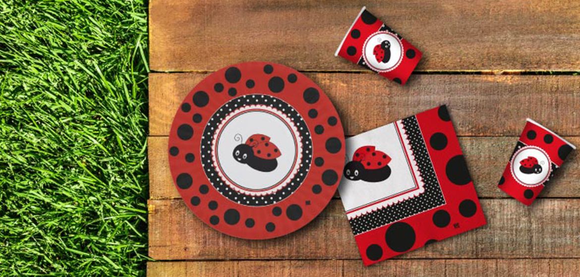 Set di articoli da festa con motivo di coccinelle rosse e nere, comprendente piatti, bicchieri e tovaglioli decorati, su uno sfondo di legno e erba verde.