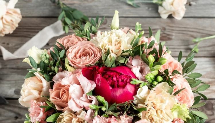 Un bouquet di fiori freschi con rose rosa, garofani bianchi e una peonia rossa, arricchito da foglie verdi, su uno sfondo di legno rustico.