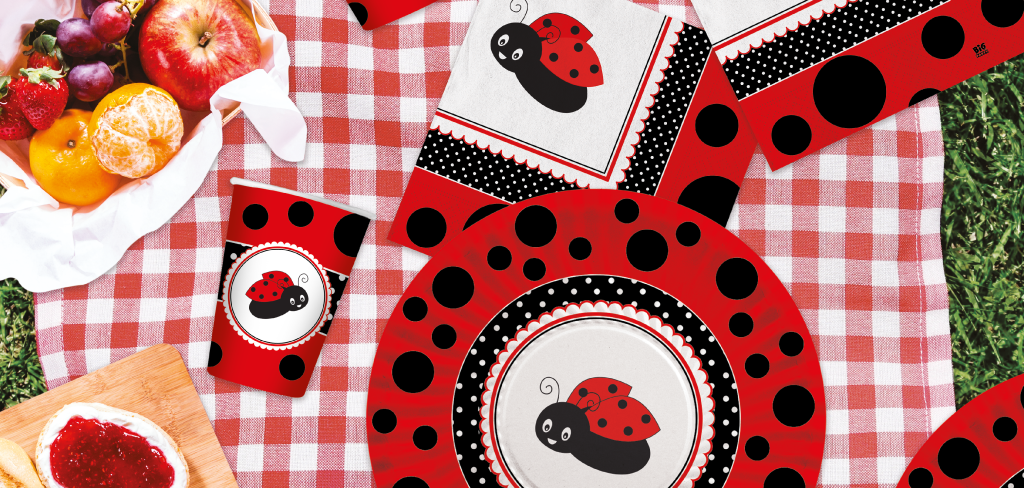 Set di articoli da festa a tema coccinella, con piatti, bicchieri e tovaglioli decorati con motivi di coccinelle rosse e nere, disposti su una tovaglia a quadri rossi e bianchi, accompagnati da frutta fresca.