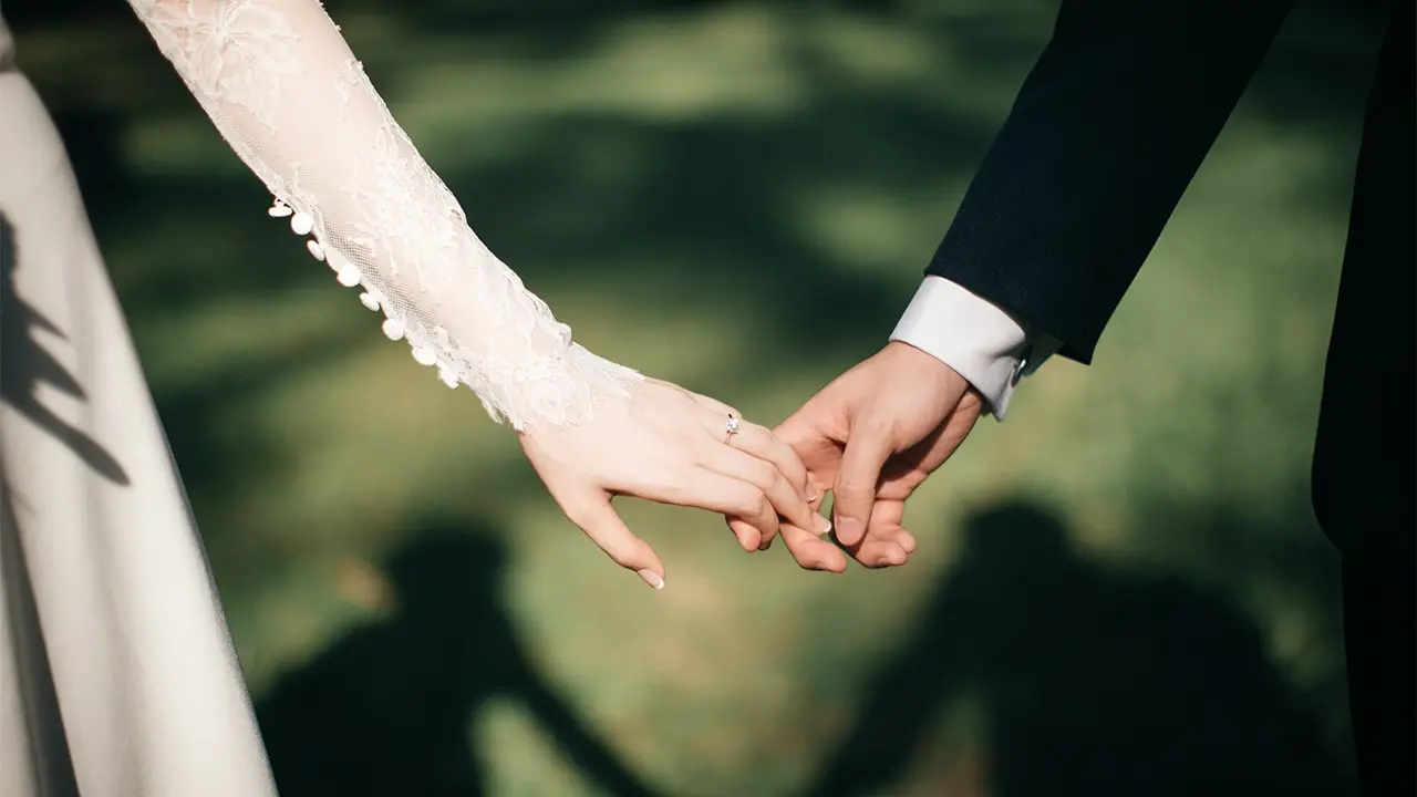 Le mani di una coppia di sposi che si tengono, con l'anello di matrimonio visibile sulla mano della sposa, sullo sfondo di un paesaggio verde.